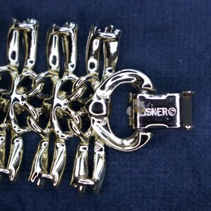 May include: Close-up of a gold-toned bracelet with a unique linked design. The bracelet features a series of stylized, interconnected figures. The clasp is rectangular and marked with the brand name "LISNER" on a dark blue background.