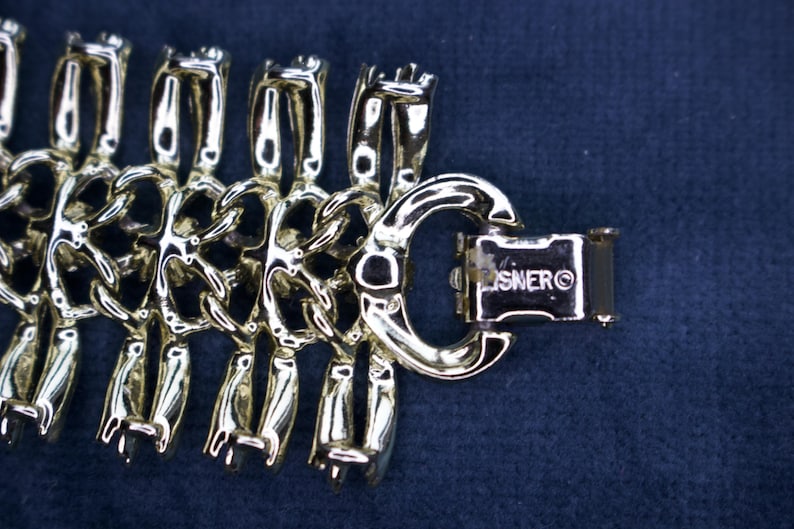 May include: Close-up of a gold-toned bracelet with a unique linked design. The bracelet features a series of stylized, interconnected figures. The clasp is rectangular and marked with the brand name "LISNER" on a dark blue background.