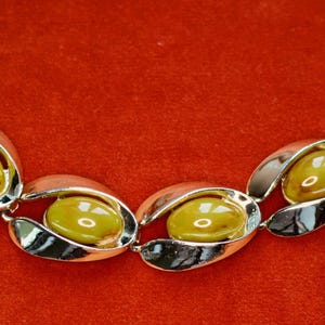 May include: A vintage bracelet with oval-shaped links. Each link features a silver-toned metal frame holding a yellow-green, oval-shaped stone. The bracelet is displayed on a red surface.