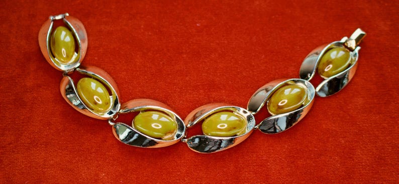 May include: A vintage bracelet with oval-shaped links. Each link features a silver-toned metal frame holding a yellow-green, oval-shaped stone. The bracelet is displayed on a red surface.