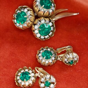 Puede incluir: Juego de joyas vintage con un broche y dos pares de pendientes. Las piezas tienen un engaste de metal dorado con una piedra central verde esmeralda rodeada de peque&ntilde;os cristales transparentes. Las joyas se exhiben sobre una superficie de terciopelo rojo.