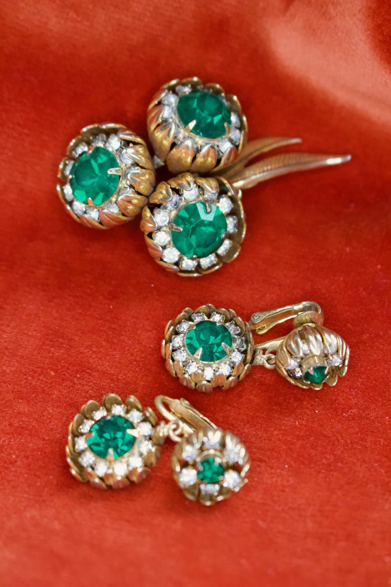 Puede incluir: Juego de joyas vintage con un broche y dos pares de pendientes. Las piezas tienen un engaste de metal dorado con una piedra central verde esmeralda rodeada de peque&ntilde;os cristales transparentes. Las joyas se exhiben sobre una superficie de terciopelo rojo.
