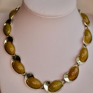 May include: A vintage necklace featuring olive-green oval-shaped stones set in silver-tone metal. The necklace is displayed on a white mannequin bust. The stones have a glossy finish, and the metal has a polished appearance.