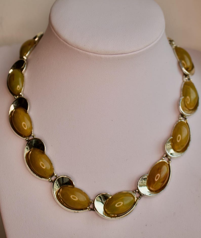 May include: A vintage necklace featuring olive-green oval-shaped stones set in silver-tone metal. The necklace is displayed on a white mannequin bust. The stones have a glossy finish, and the metal has a polished appearance.