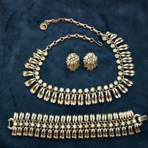 May include: A gold-toned jewelry set featuring a necklace, earrings, and bracelet. The necklace and bracelet have a repeating design of vertical bars and small, round, clear stones. The earrings are round with a cluster of clear stones.