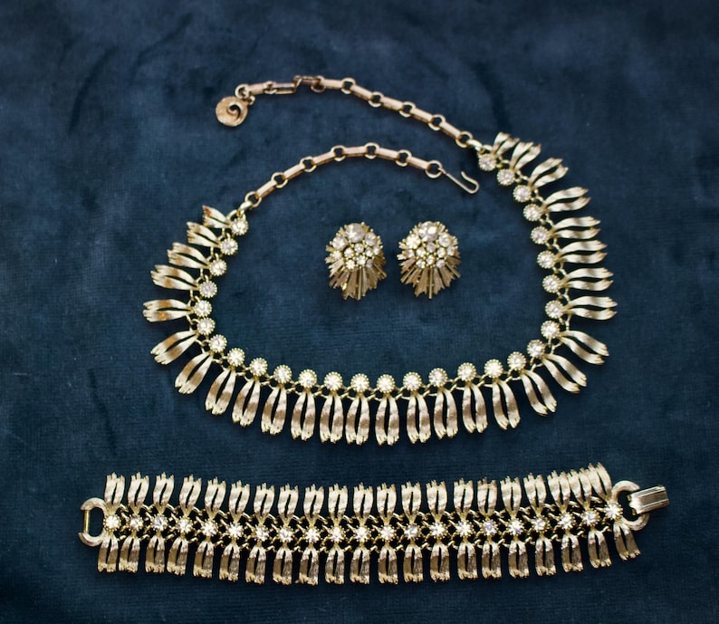 May include: A gold-toned jewelry set featuring a necklace, earrings, and bracelet. The necklace and bracelet have a repeating design of vertical bars and small, round, clear stones. The earrings are round with a cluster of clear stones.
