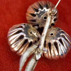 Puede incluir: Broche vintage con dise&ntilde;o floral. El broche presenta tres elementos estilizados en forma de flor calada en tono dorado, con un tallo y un alfiler plateados. El fondo es de color rojo liso.