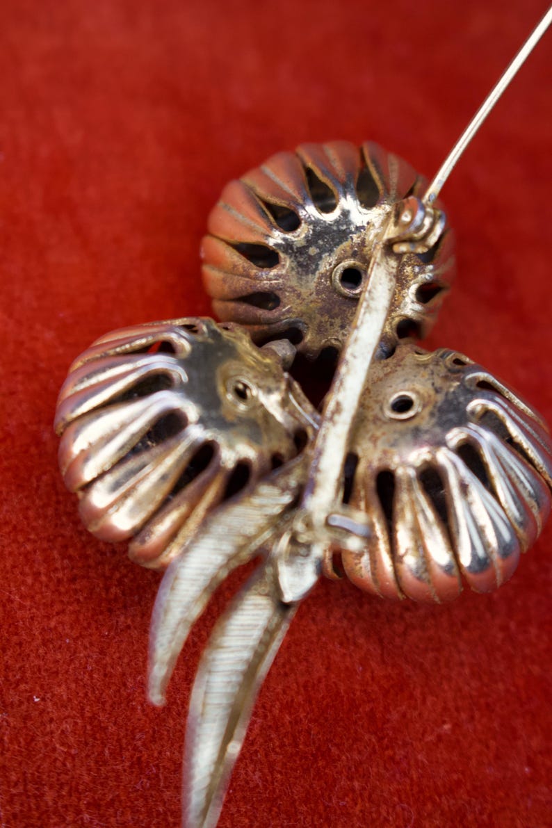 Puede incluir: Broche vintage con dise&ntilde;o floral. El broche presenta tres elementos estilizados en forma de flor calada en tono dorado, con un tallo y un alfiler plateados. El fondo es de color rojo liso.