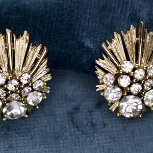 May include: A pair of gold-toned, clip-on earrings. Each earring features a sunburst design with radiating lines and is adorned with multiple clear, round, and sparkling crystals. The earrings are set against a dark teal background.