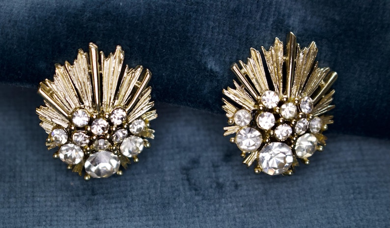 May include: A pair of gold-toned, clip-on earrings. Each earring features a sunburst design with radiating lines and is adorned with multiple clear, round, and sparkling crystals. The earrings are set against a dark teal background.