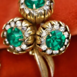 Puede incluir: Broche floral dorado con tres racimos en forma de flor. Cada racimo presenta una gran gema verde esmeralda facetada rodeada de peque&ntilde;os cristales transparentes. El broche tiene un dise&ntilde;o detallado y texturizado sobre un fondo rojo.