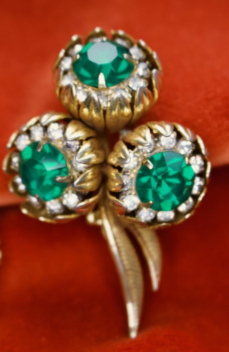 Puede incluir: Broche floral dorado con tres racimos en forma de flor. Cada racimo presenta una gran gema verde esmeralda facetada rodeada de peque&ntilde;os cristales transparentes. El broche tiene un dise&ntilde;o detallado y texturizado sobre un fondo rojo.