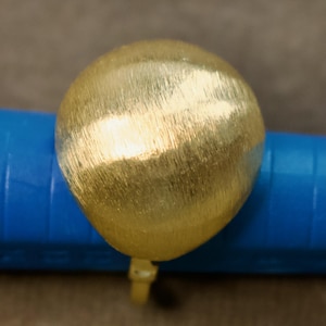 Può includere: Un anello color oro con una grande parte superiore rotonda e strutturata. L'anello è posto su una superficie blu. Il design dell'anello presenta una finitura in metallo spazzolato, che gli conferisce un aspetto unico e moderno. È un gioiello.