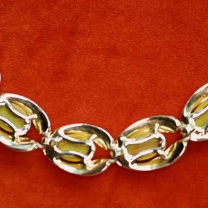 May include: A vintage bracelet featuring oval-shaped links with a gold-tone metal frame and a pale yellow center. The bracelet has a clasp closure and is displayed against a red background.