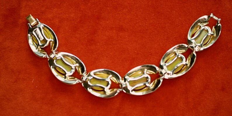 May include: A vintage bracelet featuring oval-shaped links with a gold-tone metal frame and a pale yellow center. The bracelet has a clasp closure and is displayed against a red background.