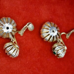 Puede incluir: Un par de pendientes vintage con un dise&ntilde;o floral. Los pendientes presentan una parte superior plateada en forma de flor con un borde dorado y una peque&ntilde;a flor colgante. Sobre un fondo rojo.