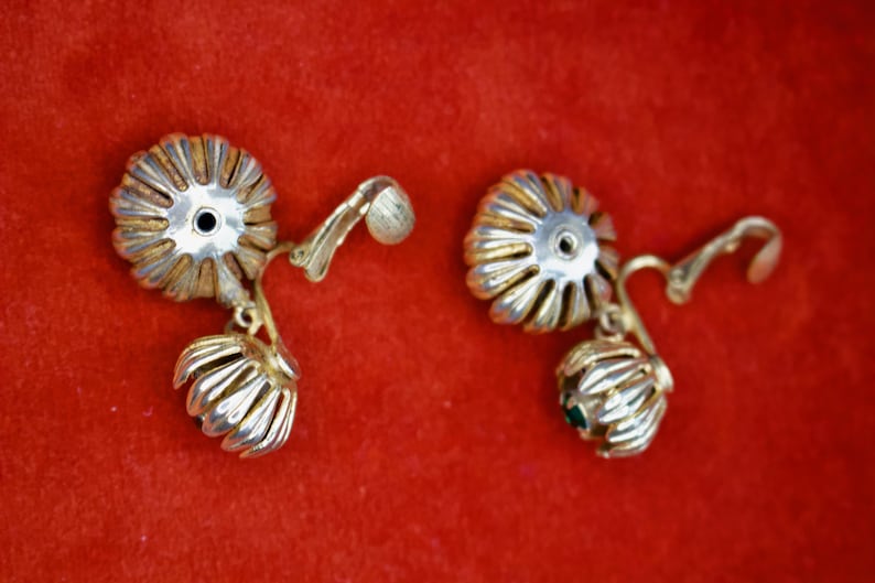 Puede incluir: Un par de pendientes vintage con un dise&ntilde;o floral. Los pendientes presentan una parte superior plateada en forma de flor con un borde dorado y una peque&ntilde;a flor colgante. Sobre un fondo rojo.