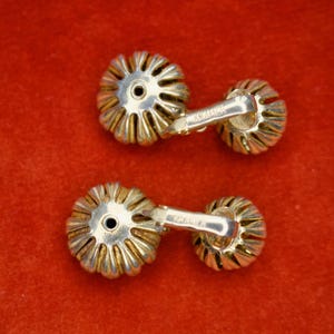 Puede incluir: Un par de gemelos vintage con un dise&ntilde;o floral. Los gemelos tienen una base de metal plateado con detalles dorados. El dise&ntilde;o es una serie de p&eacute;talos que irradian desde un punto central. Los gemelos est&aacute;n sobre un fondo rojo.
