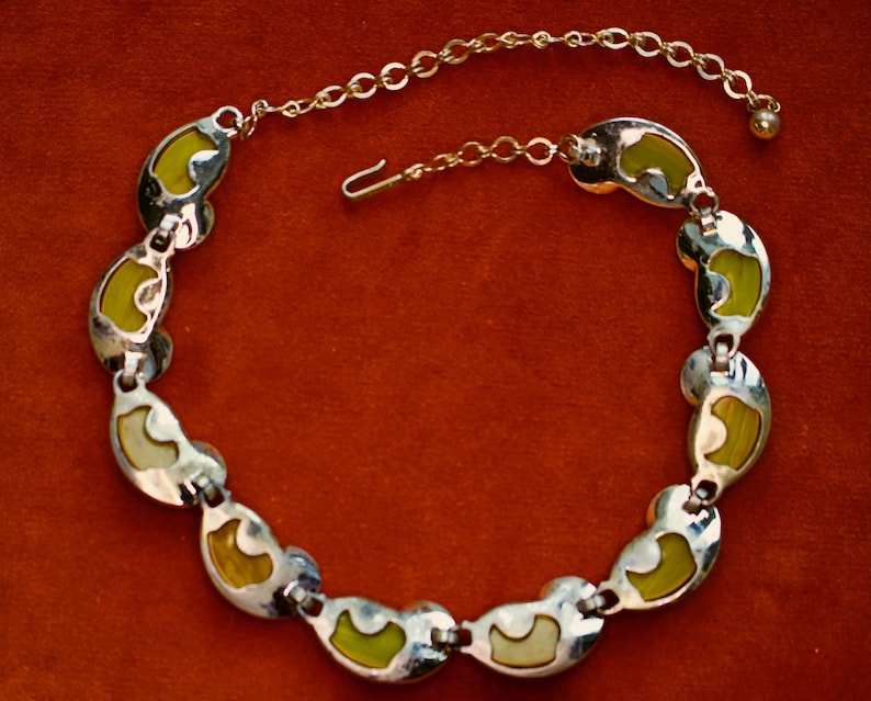 May include: A vintage necklace with a silver-tone metal chain and links. Each link features a kidney-shaped design with a lime green and cream-colored enamel inlay. The necklace has a hook clasp and an extender chain.