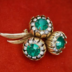 Puede incluir: Broche dorado con tres racimos en forma de flor, cada uno con una gran piedra central verde esmeralda rodeada de peque&ntilde;os cristales transparentes. El broche tiene un dise&ntilde;o de hoja que se extiende desde las flores, sobre un fondo rojo.