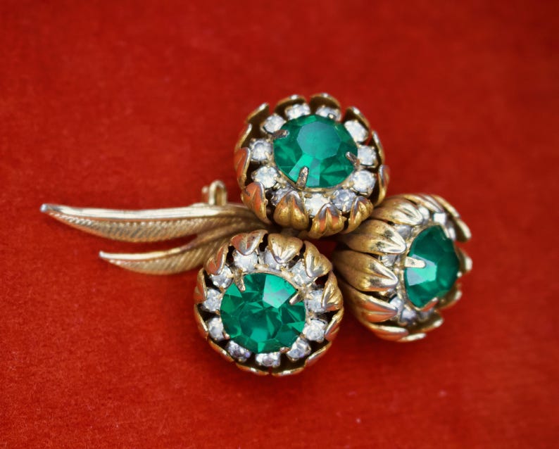 Puede incluir: Broche dorado con tres racimos en forma de flor, cada uno con una gran piedra central verde esmeralda rodeada de peque&ntilde;os cristales transparentes. El broche tiene un dise&ntilde;o de hoja que se extiende desde las flores, sobre un fondo rojo.
