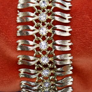 May include: Gold-toned bracelet with a ribbon design, featuring clear, round gemstones at intervals. The bracelet is set against a red background, highlighting its metallic sheen and the sparkle of the stones. A vintage jewelry piece.