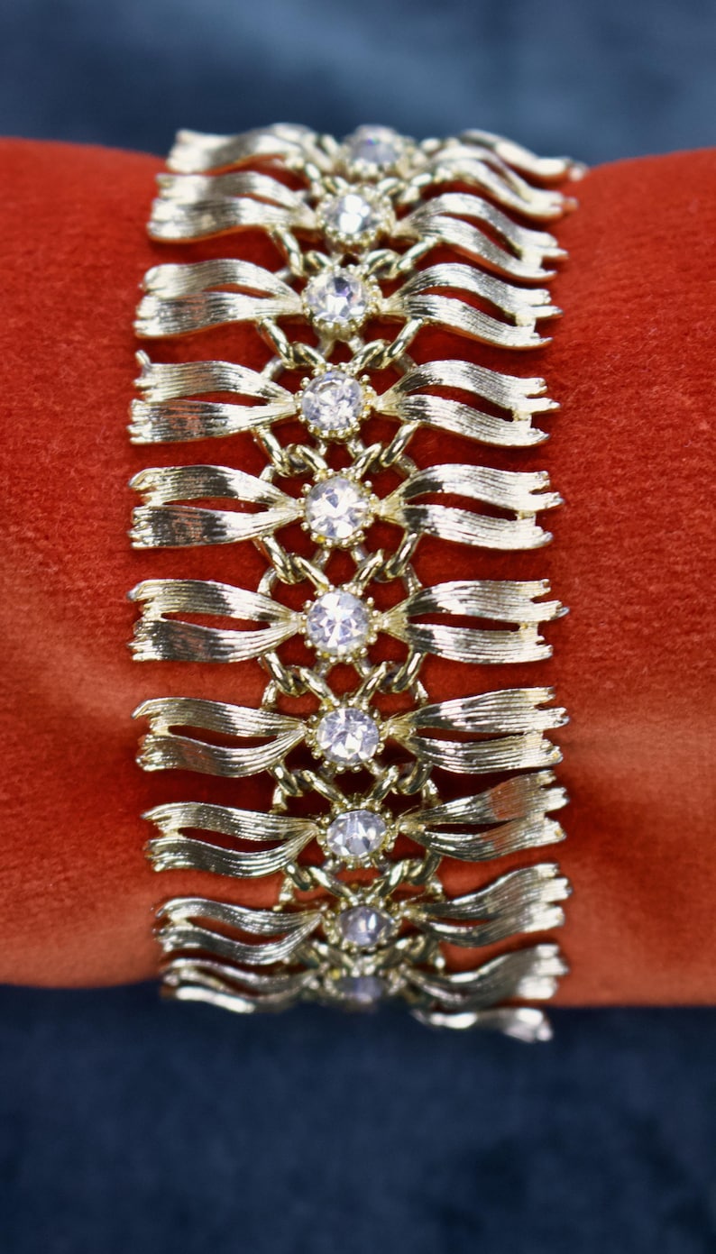 May include: Gold-toned bracelet with a ribbon design, featuring clear, round gemstones at intervals. The bracelet is set against a red background, highlighting its metallic sheen and the sparkle of the stones. A vintage jewelry piece.