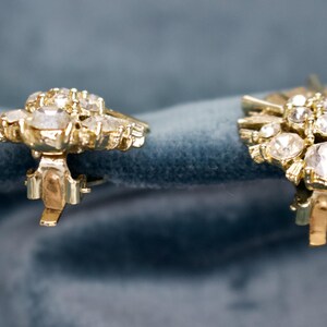 May include: A pair of vintage gold-toned clip-on earrings. Each earring features a cluster of clear, sparkling rhinestones set in a floral design. The earrings are displayed against a blue velvet background.