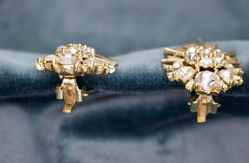 May include: A pair of vintage gold-toned clip-on earrings. Each earring features a cluster of clear, sparkling rhinestones set in a floral design. The earrings are displayed against a blue velvet background.