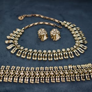 May include: A gold-toned jewelry set featuring a necklace, bracelet, and matching earrings. The necklace and bracelet have a repeating design of curved, ribbon-like elements with small, clear gemstones. The earrings are round with a textured, floral-like design.