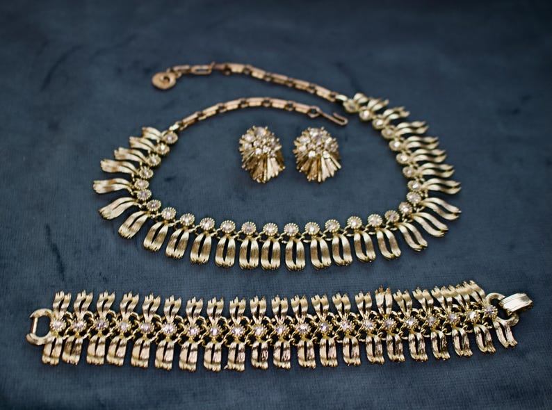 May include: A gold-toned jewelry set featuring a necklace, bracelet, and matching earrings. The necklace and bracelet have a repeating design of curved, ribbon-like elements with small, clear gemstones. The earrings are round with a textured, floral-like design.