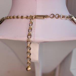 May include: A gold-toned necklace with an adjustable chain and clasp. The necklace is displayed on a white mannequin. The chain has a small ball at the end of the adjustable portion.