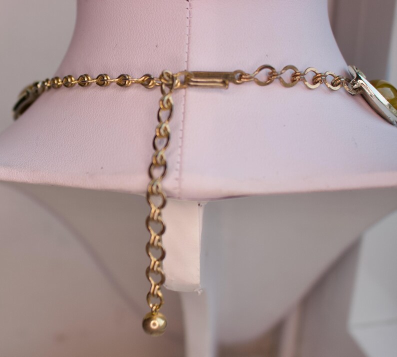May include: A gold-toned necklace with an adjustable chain and clasp. The necklace is displayed on a white mannequin. The chain has a small ball at the end of the adjustable portion.