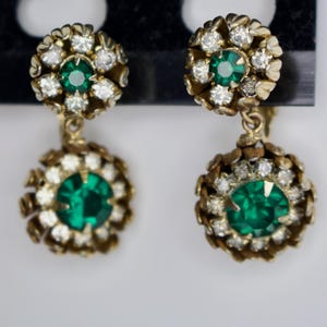 Puede incluir: Un par de pendientes vintage dorados. Cada pendiente presenta un grupo de peque&ntilde;os cristales transparentes que rodean una piedra central redonda de color verde esmeralda. Los pendientes tienen un dise&ntilde;o colgante con una piedra verde esmeralda similar y m&aacute;s grande en la parte inferior.