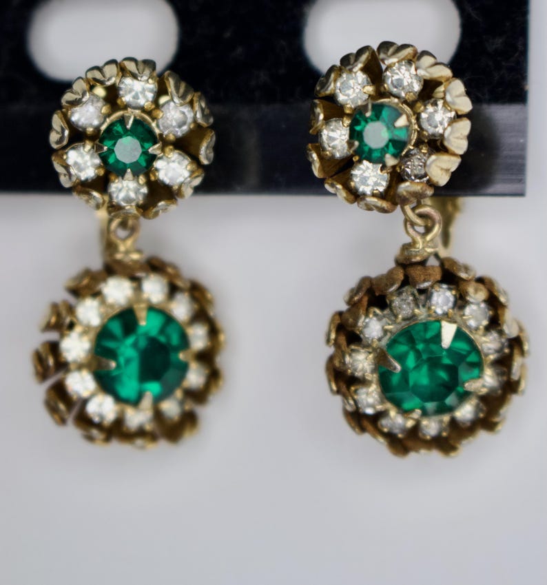 Puede incluir: Un par de pendientes vintage dorados. Cada pendiente presenta un grupo de peque&ntilde;os cristales transparentes que rodean una piedra central redonda de color verde esmeralda. Los pendientes tienen un dise&ntilde;o colgante con una piedra verde esmeralda similar y m&aacute;s grande en la parte inferior.