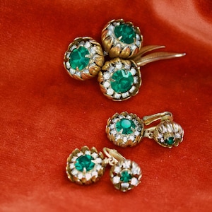 Könnte beinhalten: Eine Sammlung von Vintage-Schmuckstücken, darunter eine Brosche und Ohrringe, mit goldfarbenem Metall und leuchtend grünen Edelsteinen. Die Edelsteine sind von kleinen, klaren Steinen umgeben, die auf rotem Hintergrund funkeln.