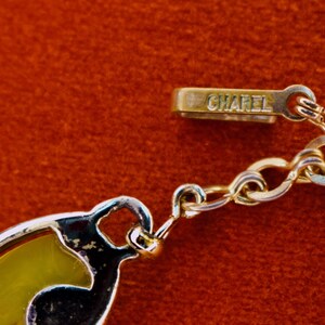 May include: Close-up of a gold-toned necklace with a decorative pendant. The necklace features a chain and a clasp with the word "CHAREL" engraved. The pendant has a yellow and black design, set against a red background.