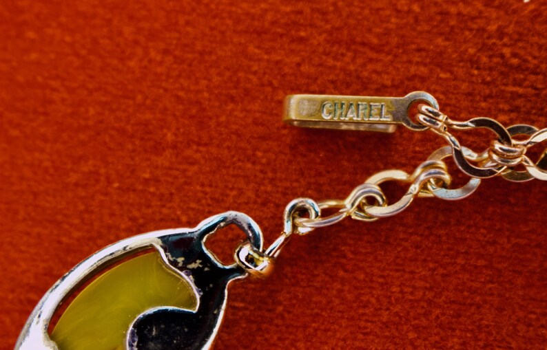 May include: Close-up of a gold-toned necklace with a decorative pendant. The necklace features a chain and a clasp with the word "CHAREL" engraved. The pendant has a yellow and black design, set against a red background.