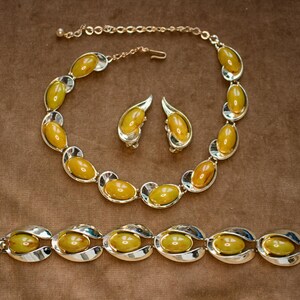 May include: A vintage jewelry set featuring a necklace, earrings, and bracelet. The set has oval-shaped amber-colored stones set in silver-tone metal. The necklace and bracelet have a curved design, and the earrings are clip-on style.