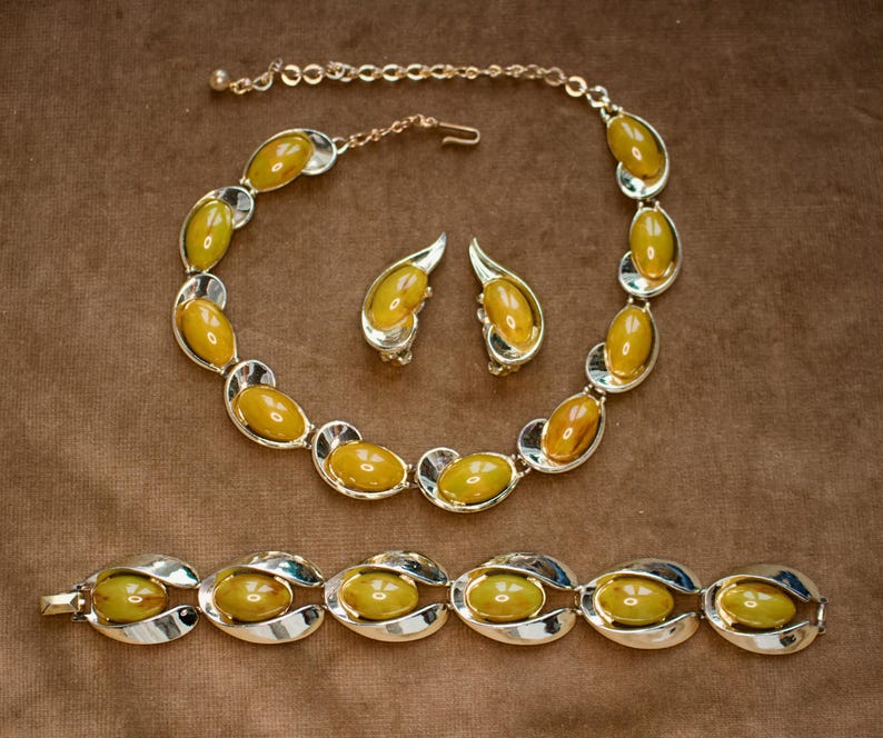 May include: A vintage jewelry set featuring a necklace, earrings, and bracelet. The set has oval-shaped amber-colored stones set in silver-tone metal. The necklace and bracelet have a curved design, and the earrings are clip-on style.