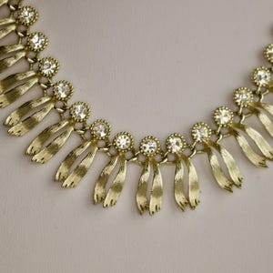 May include: A gold-toned necklace with a series of stylized ribbon-like pendants. Each pendant is connected by a small, round, clear rhinestone. The necklace is displayed on a white surface.