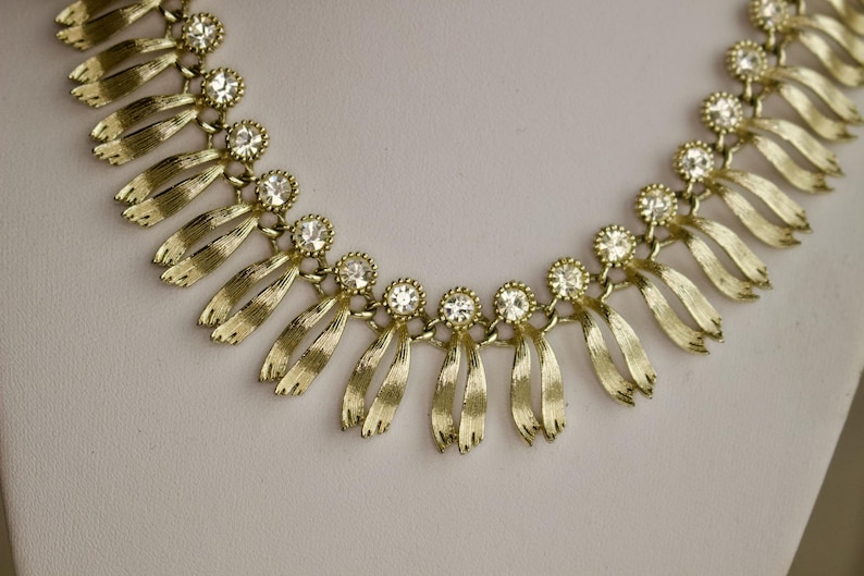 May include: A gold-toned necklace with a series of stylized ribbon-like pendants. Each pendant is connected by a small, round, clear rhinestone. The necklace is displayed on a white surface.