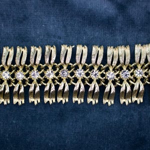 May include: Gold-toned bracelet with a repeating pattern of stylized bows and clear, round gemstones. The bracelet is set against a dark blue background, highlighting its metallic sheen and intricate design. A vintage-inspired piece.