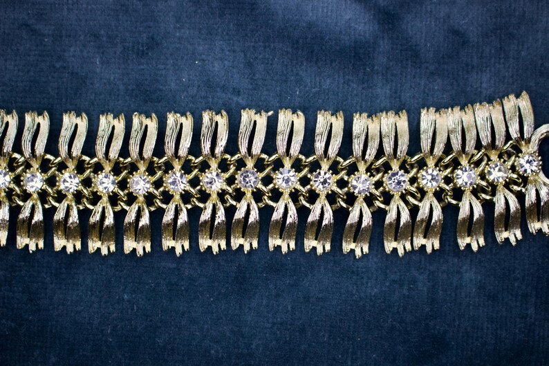 May include: Gold-toned bracelet with a repeating pattern of stylized bows and clear, round gemstones. The bracelet is set against a dark blue background, highlighting its metallic sheen and intricate design. A vintage-inspired piece.