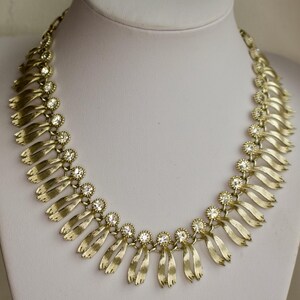 May include: Gold-toned necklace with a series of stylized ribbon-like pendants. Each pendant is accented with a cluster of clear, round rhinestones. The necklace is displayed on a white mannequin bust.