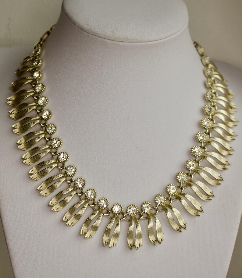 May include: Gold-toned necklace with a series of stylized ribbon-like pendants. Each pendant is accented with a cluster of clear, round rhinestones. The necklace is displayed on a white mannequin bust.