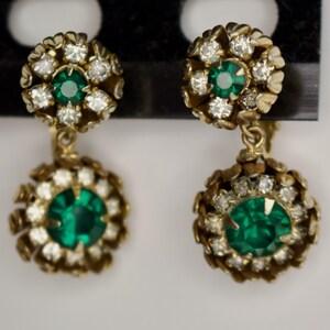 Puede incluir: Un par de pendientes dorados vintage. Cada pendiente presenta una piedra central de color verde esmeralda rodeada de peque&ntilde;os cristales transparentes. El dise&ntilde;o incluye un grupo m&aacute;s peque&ntilde;o en la parte superior y un grupo m&aacute;s grande colgante en la parte inferior.