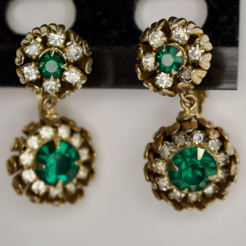 Puede incluir: Un par de pendientes dorados vintage. Cada pendiente presenta una piedra central de color verde esmeralda rodeada de peque&ntilde;os cristales transparentes. El dise&ntilde;o incluye un grupo m&aacute;s peque&ntilde;o en la parte superior y un grupo m&aacute;s grande colgante en la parte inferior.