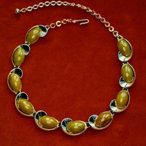 May include: A vintage necklace featuring olive-green oval-shaped stones set in silver-tone metal. The stones are framed by black accents, and the necklace has a delicate chain with a hook clasp. The background is a solid red.