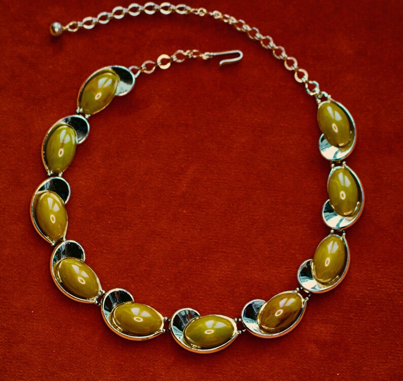 May include: A vintage necklace featuring olive-green oval-shaped stones set in silver-tone metal. The stones are framed by black accents, and the necklace has a delicate chain with a hook clasp. The background is a solid red.
